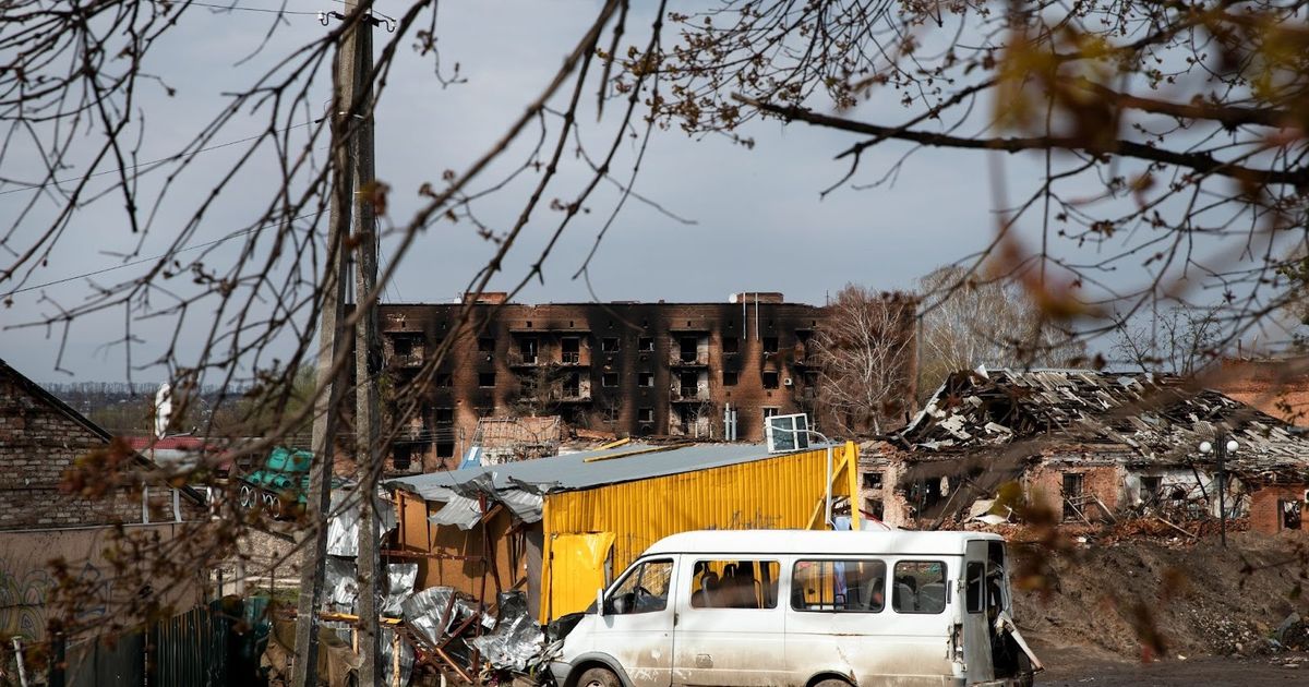Damaged residential buildings and debris in war-torn area with abandoned white van and yellow equipment structure, illustrating disaster impact on infrastructure.