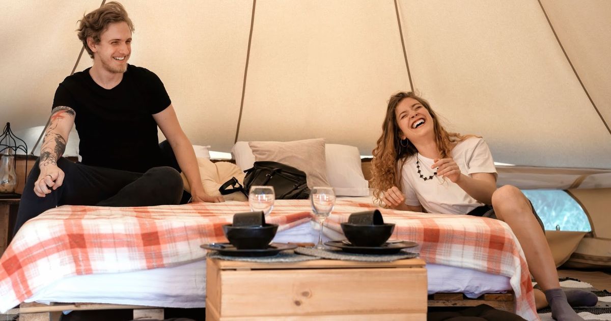 Two guests smiling inside a glamping tent with a red plaid blanket, wooden table with place settings, and water glasses visible.