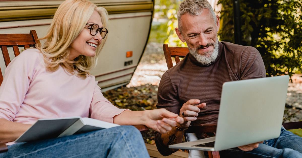 Two people reviewing a laptop together while sitting in chairs outside an RV, smiling during a campground booking discussion.