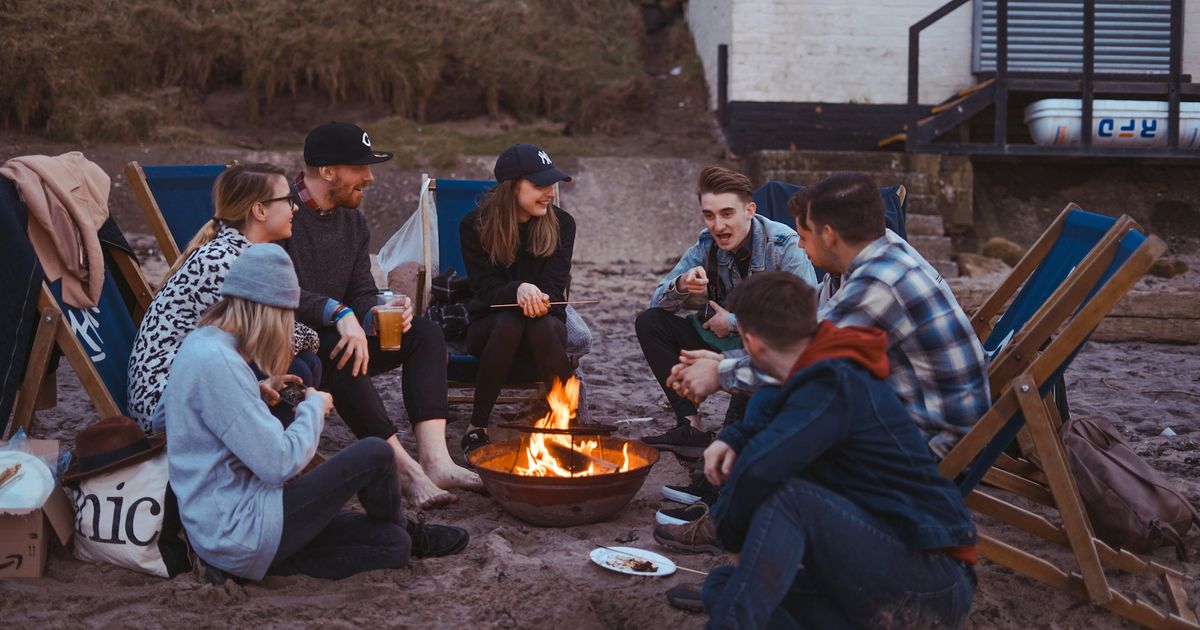 Group of friends gathered around a beach bonfire at dusk, sitting in deck chairs, socializing and enjoying drinks together.