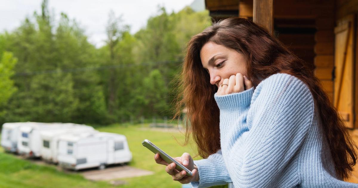 Woman in light blue sweater holding phone, looking at RV trailers in grassy campground with trees in background.
