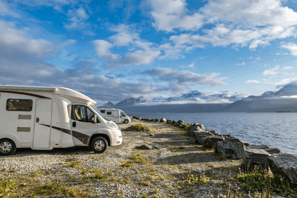 Motorhomes parked on a rocky beach overlooking snow-capped mountains and a bay under blue sky with clouds.