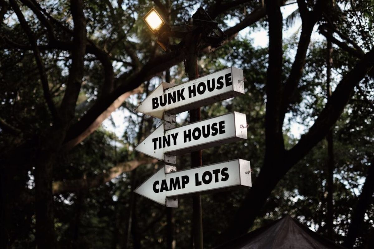 Directional signs in a wooded campground pointing to Bunk House, Tiny House, and Camp Lots, with a lit lamp above against tall trees.