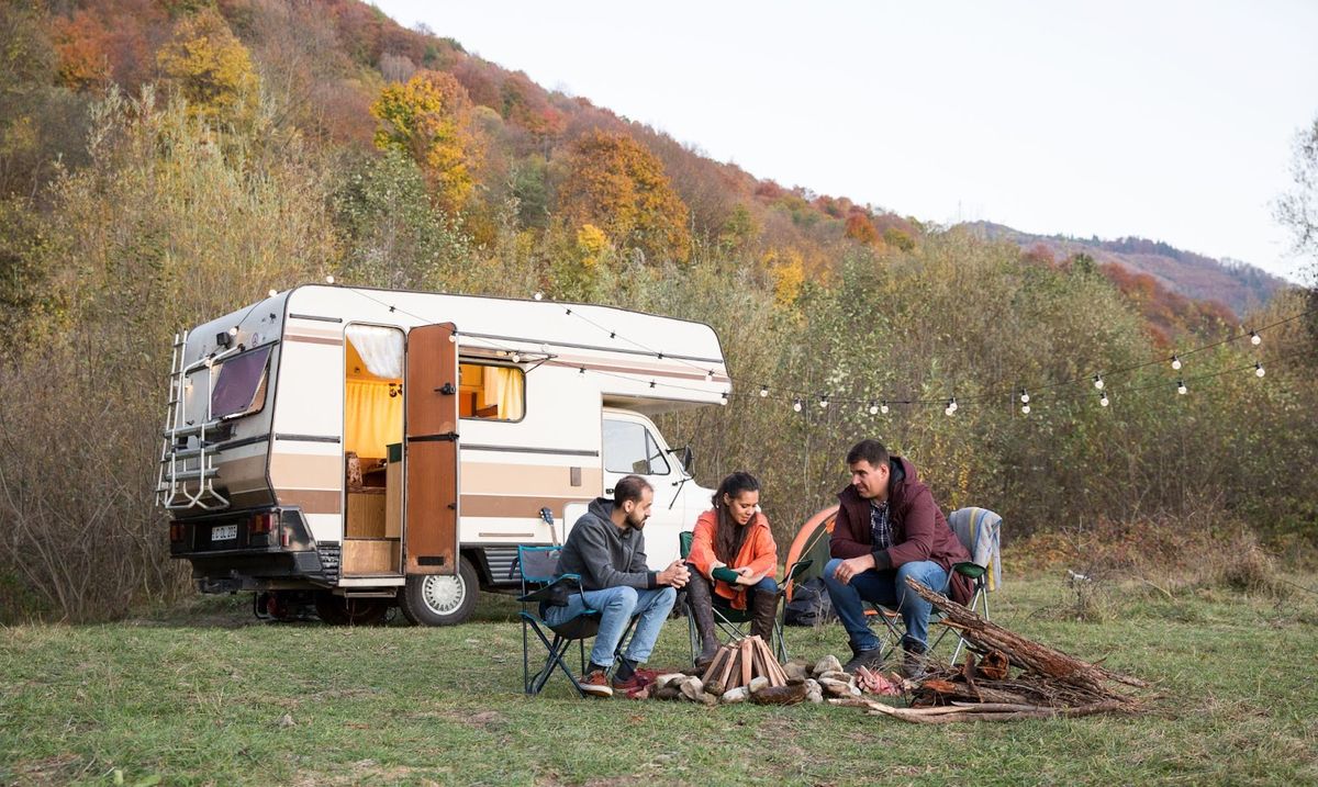 Group of four people sitting around a campfire next to a vintage RV trailer in an autumn forest landscape.