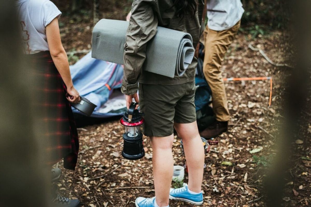 Group of campers standing outdoors with camping gear including a rolled sleeping mat and lantern on a gravel campground path.