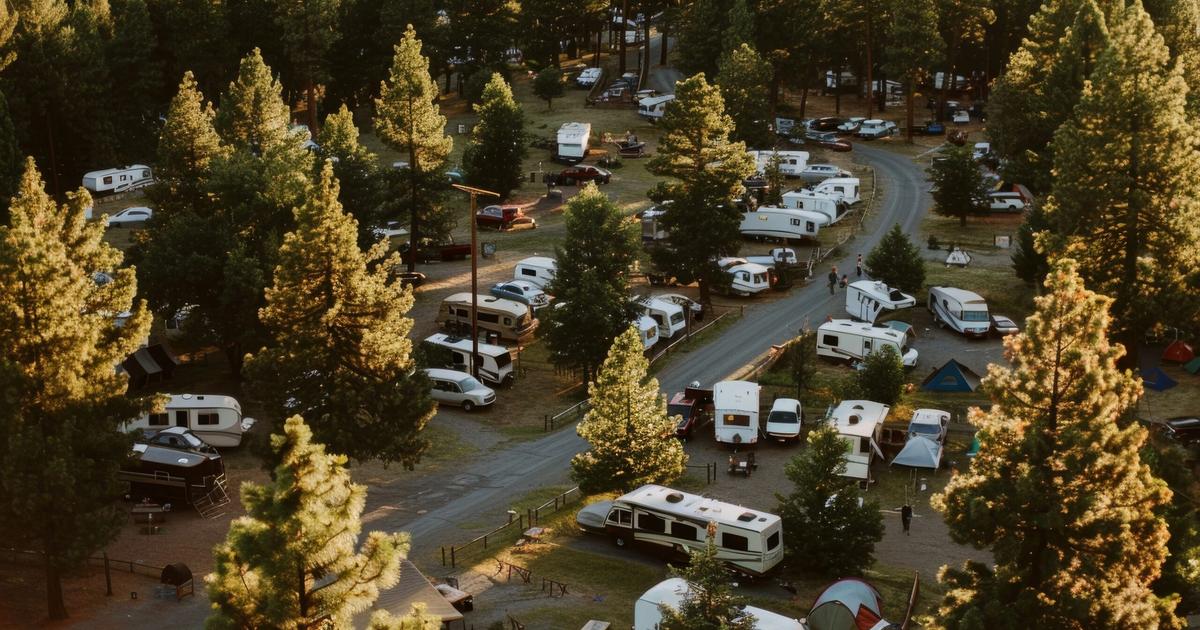 Aerial view of a campground nestled among tall pine trees with numerous RVs and trailers parked in organized sites along roads.