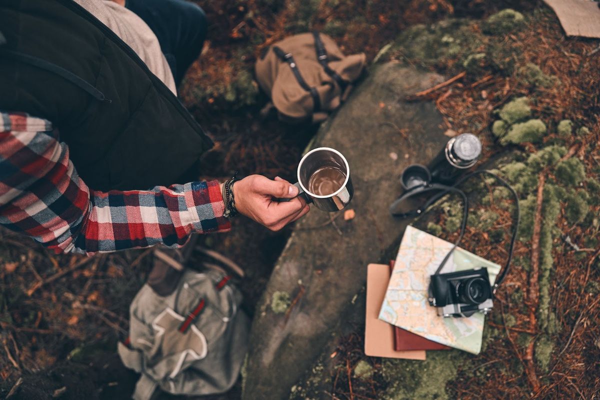 Person in plaid shirt holding metal camping mug, seated outdoors near map, camera, and notebook on mossy ground.