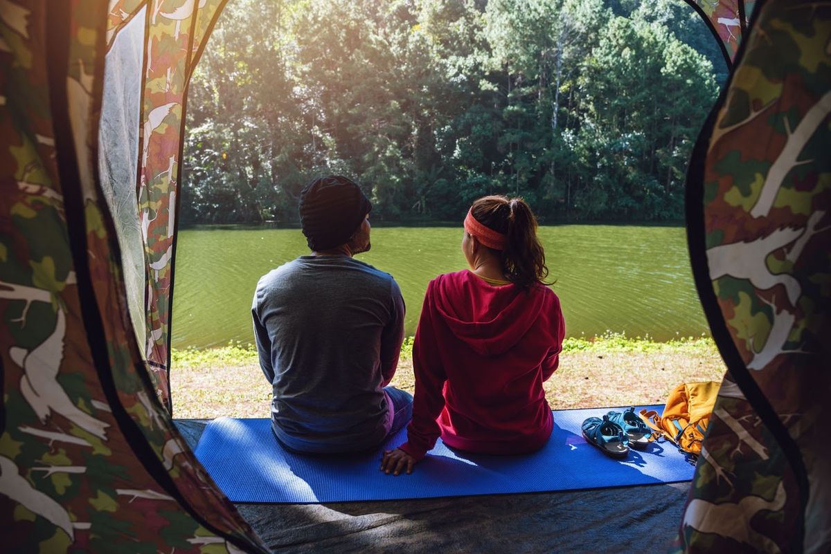 Two campers sitting inside a tent looking out at a grassy field surrounded by trees on a sunny day.