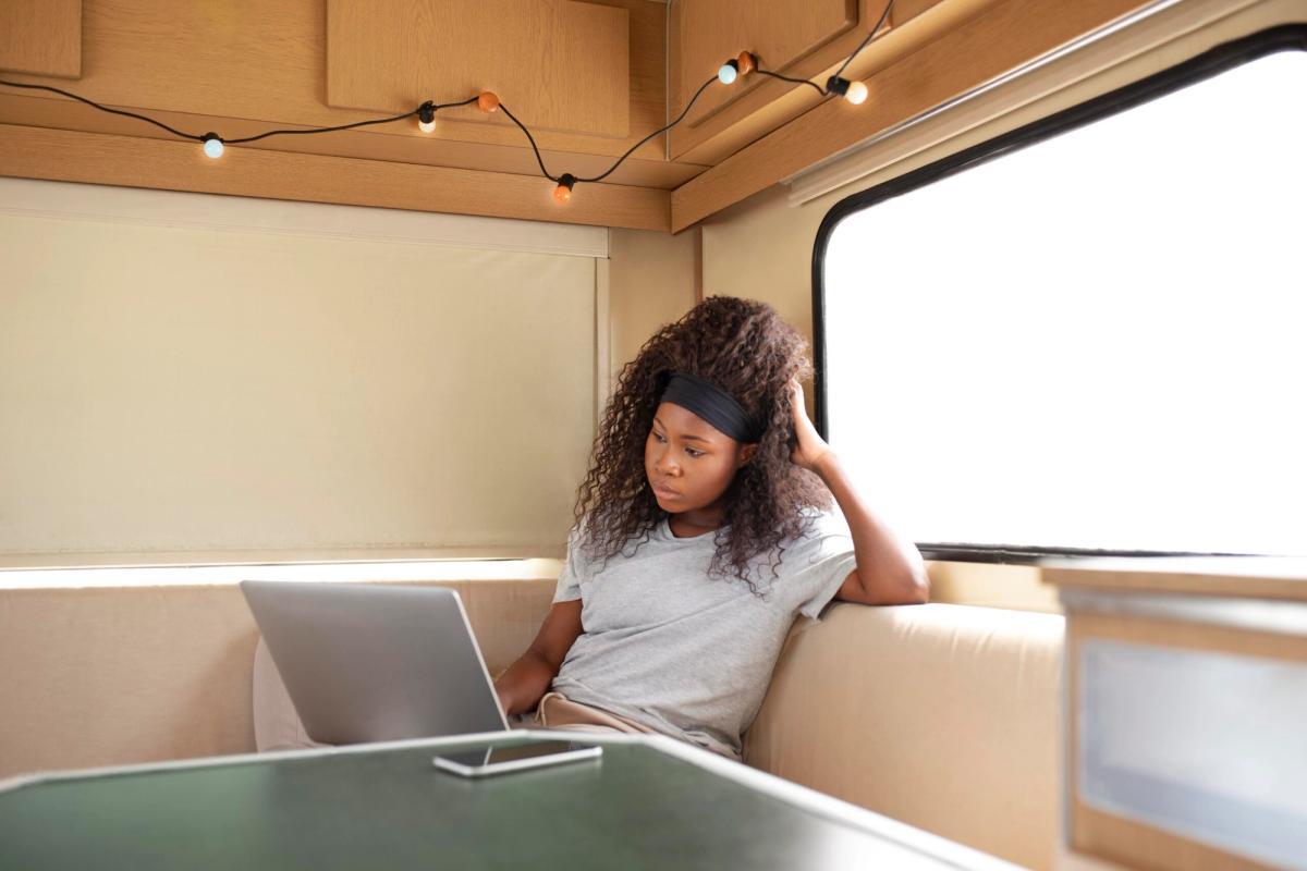 Woman working on laptop inside a wooden RV, sitting on tan upholstered seating with large window and overhead lighting.