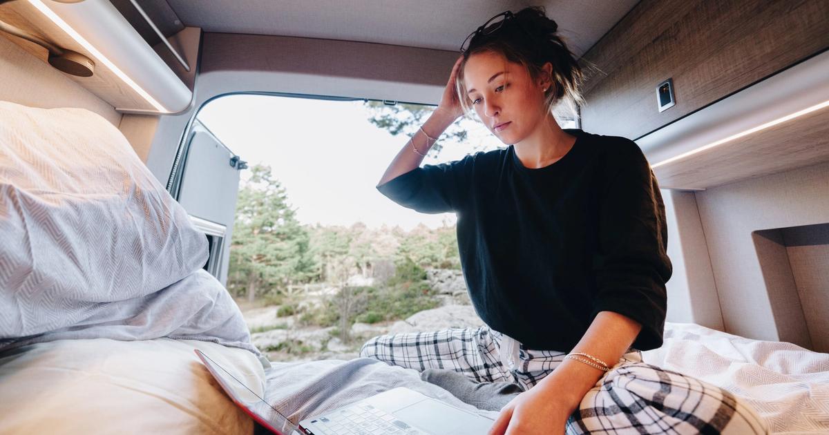 Woman sitting on RV bed reviewing map and travel plans while looking out at scenic forest landscape through window.