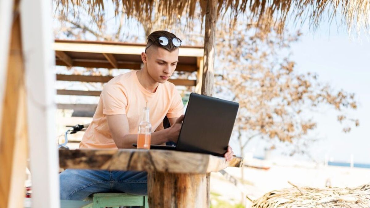 Young man working on laptop at outdoor wooden table under thatched roof, with beverage nearby and dry vegetation in background.