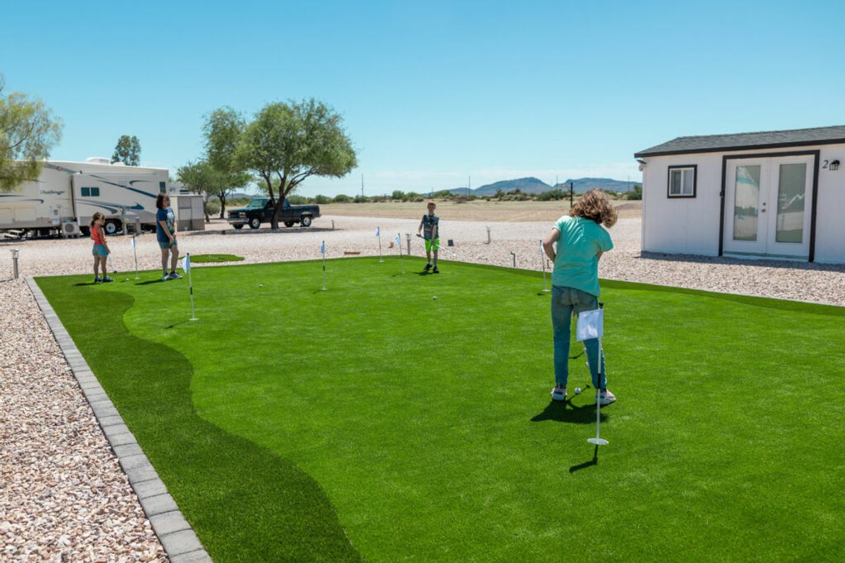 Family playing miniature golf on artificial turf putting green at RV resort with travel trailers and mountains in background