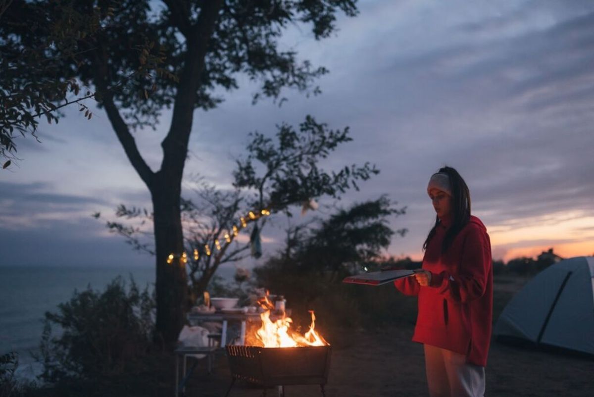 Woman in red jacket cooking at campfire during sunset, with tent and strung lights in background at mountain campsite.
