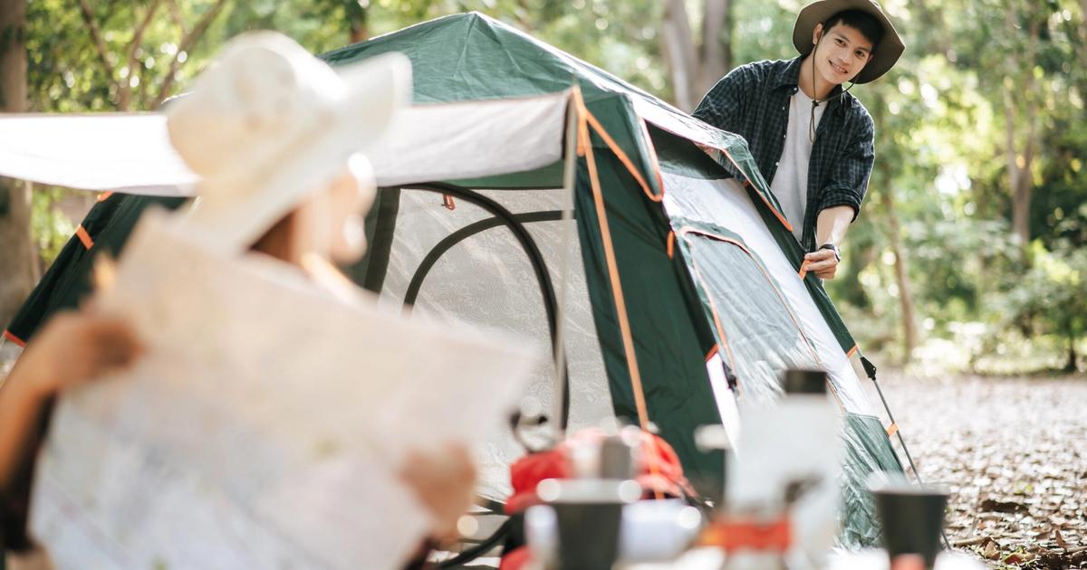 Young campers setting up a green tent in a wooded campground with camping gear and cookware spread on the ground nearby.
