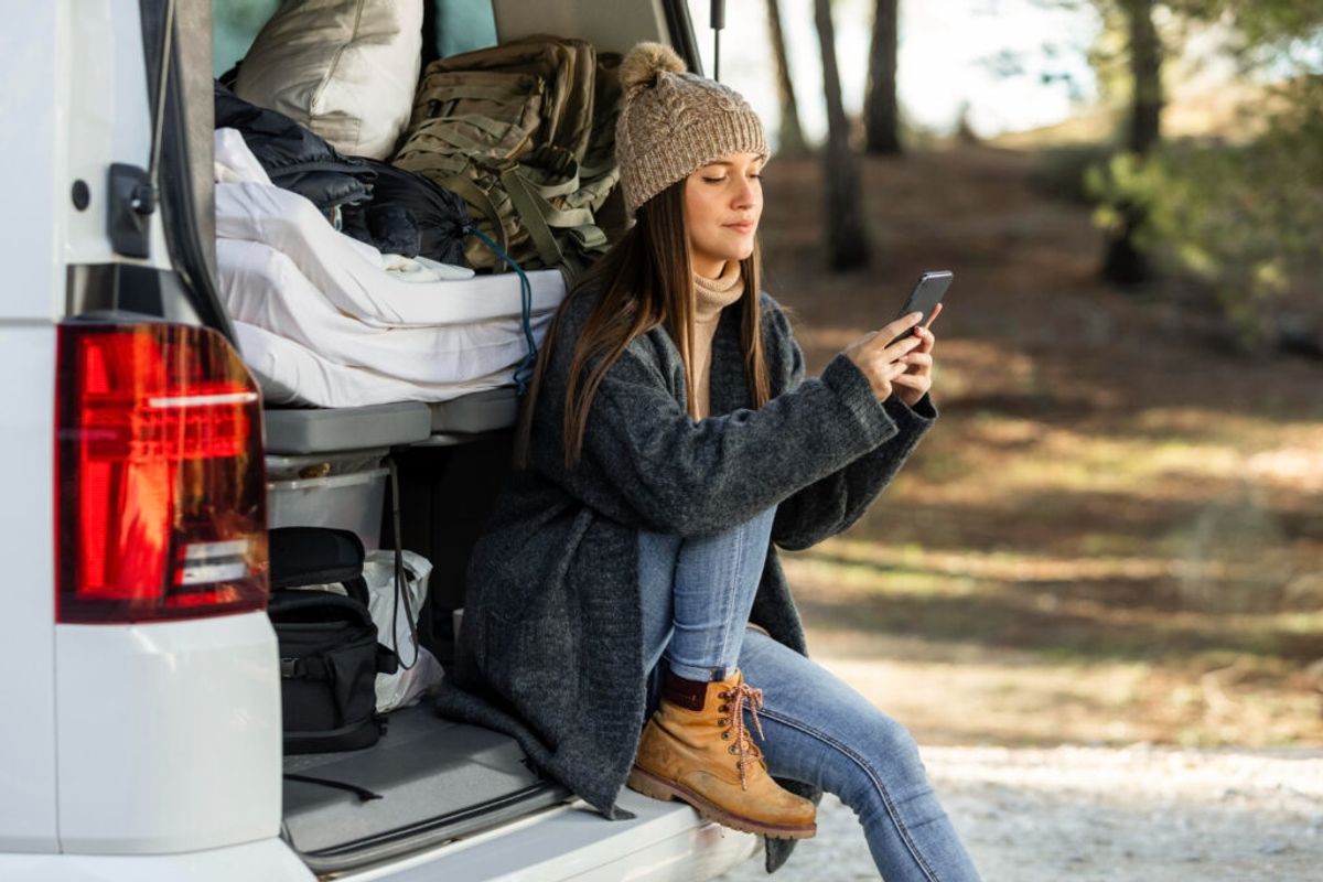 Woman sitting in open van door checking smartphone while camping in forest, surrounded by camping gear and hiking backpacks.