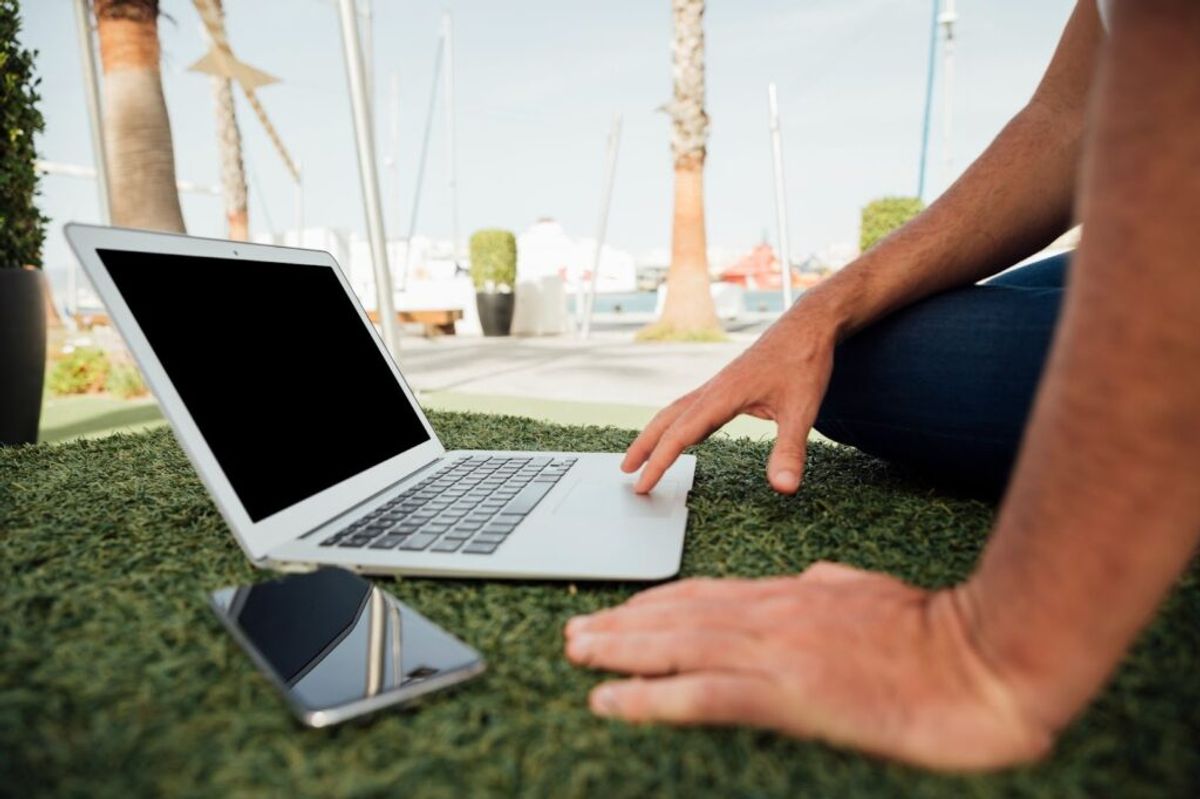 Person using laptop and smartphone on artificial grass outdoors with marina and palm trees in background