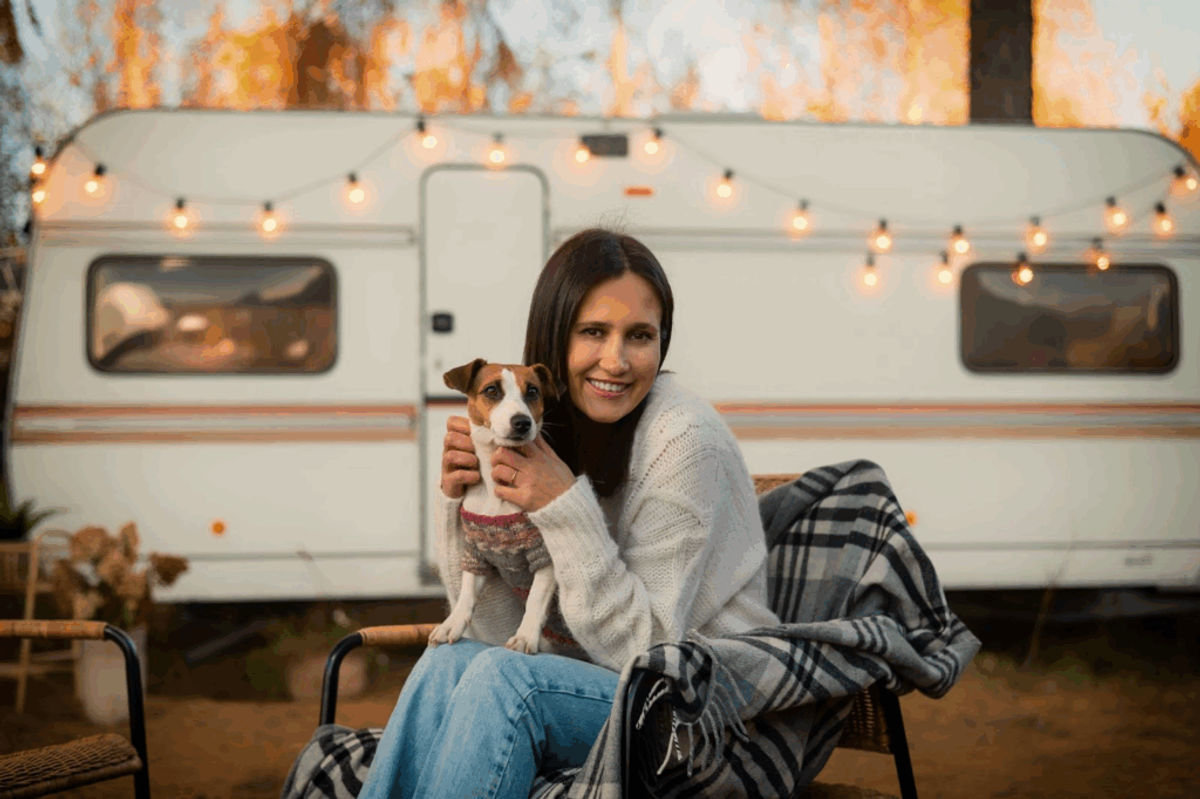 Woman holding small dog while sitting in front of vintage Airstream trailer decorated with string lights in desert setting.