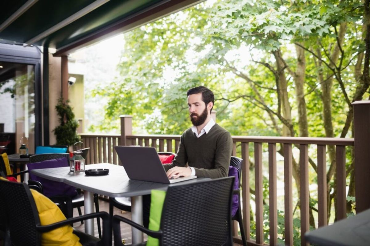 Man working on laptop at outdoor patio table with colorful chairs, overlooking forest trees and wooden deck railing.