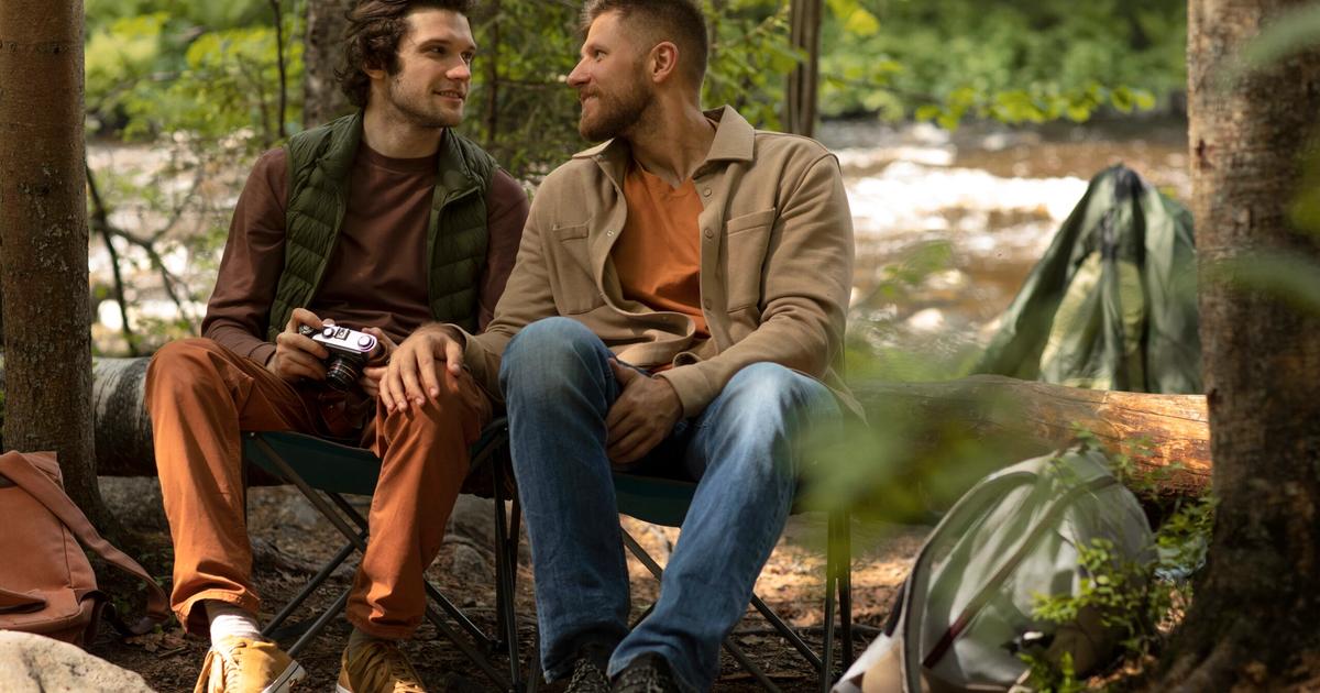Two men sitting on camping chairs in a forest campsite, smiling and conversing by a tent and river.