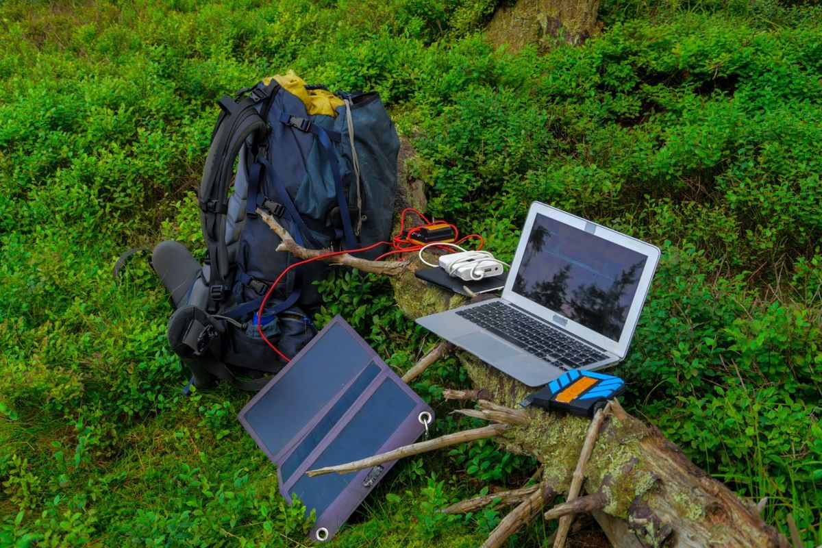 Laptop and solar panel charging setup at campsite surrounded by green moss and fallen logs, with backpack and binoculars nearby.