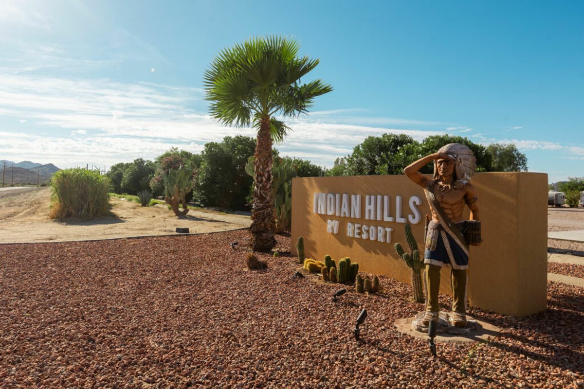 Entrance sign for Indian Hills RV Resort with Native American statue, palm tree, and desert landscape under blue sky