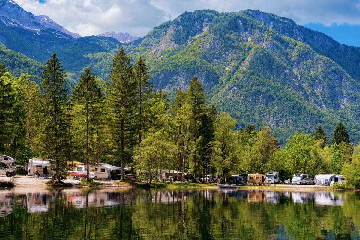 RVs and trailers parked along a scenic riverbank surrounded by forested mountains and tall pine trees under blue sky.