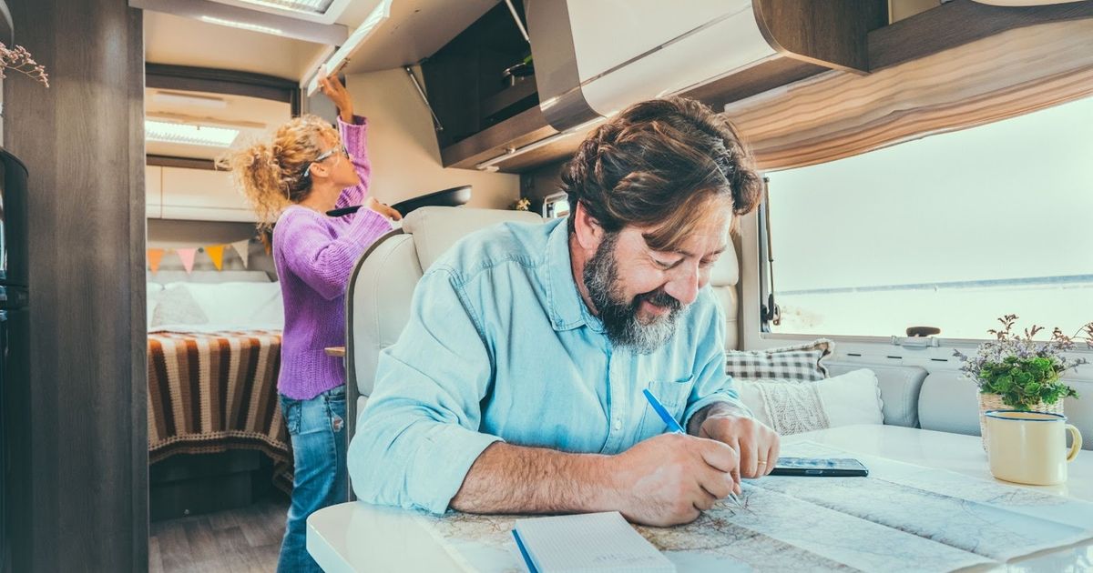 Man reviewing RV park blueprints at table inside modern motorhome while woman arranges items in kitchen cabinet above.