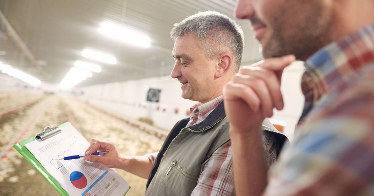 Two men reviewing a clipboard with charts and graphs inside a large greenhouse facility with rows of plants.