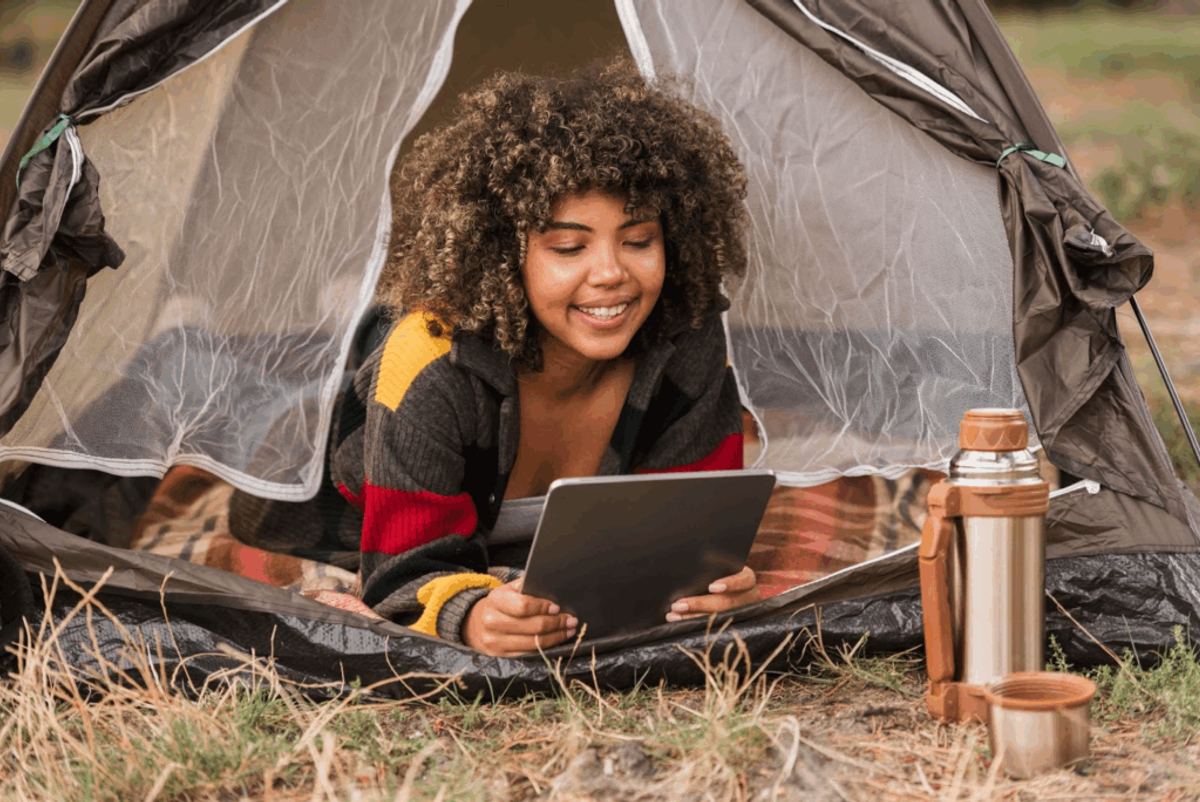 Young woman using tablet inside tent while camping, with thermos nearby on grass.