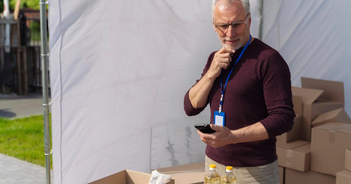 Man in maroon shirt holding phone at outdoor tent stand with bottled water and supplies on table, examining inventory.
