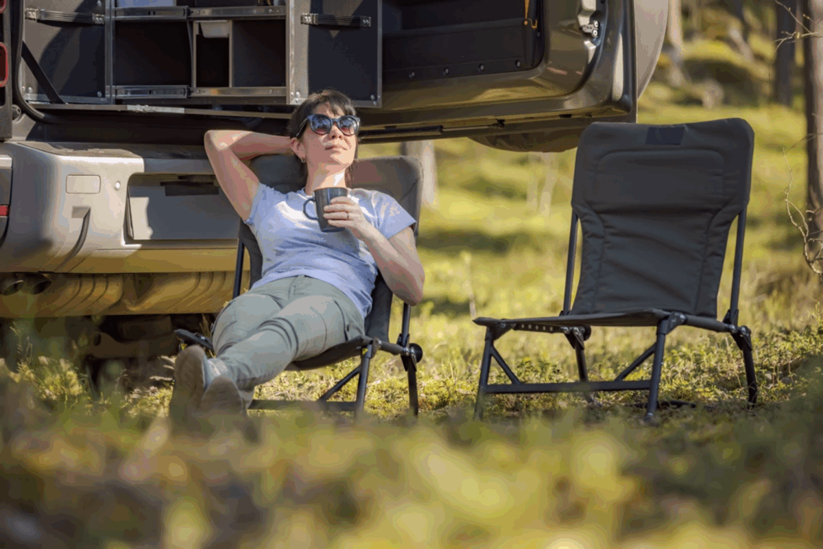Woman relaxing in a camping chair with a beverage next to her RV trailer, enjoying outdoor camping lifestyle.