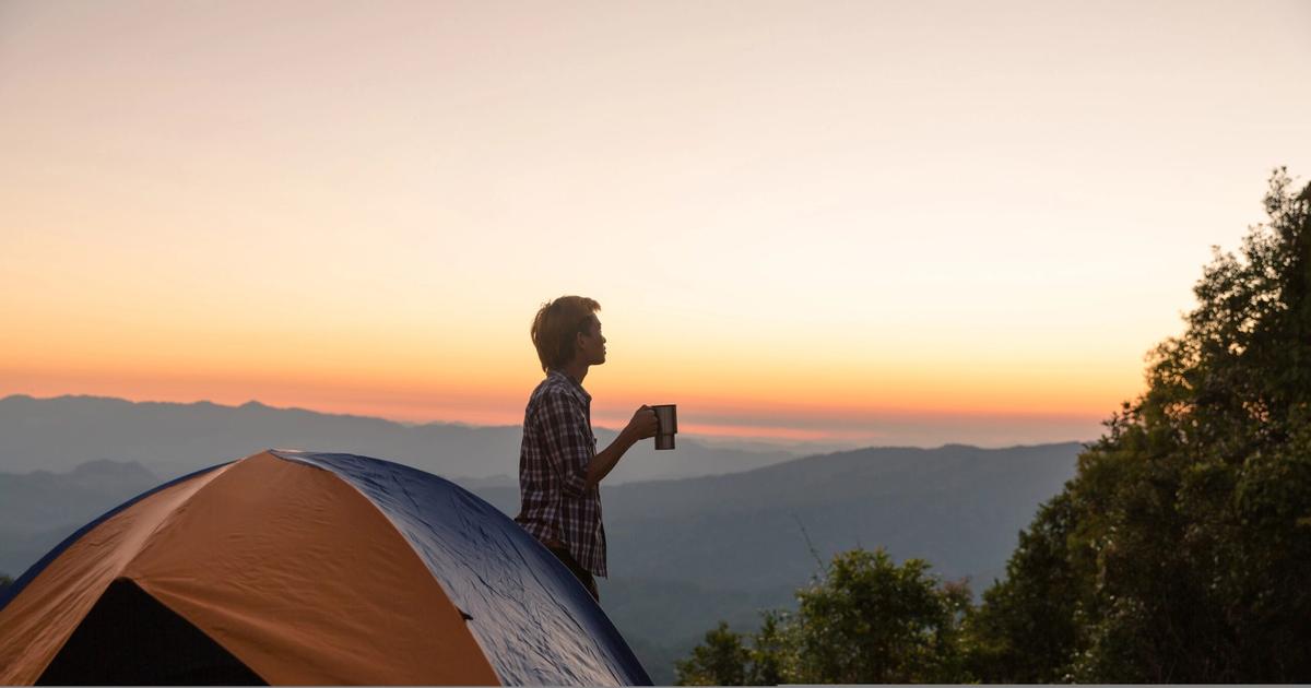 Camper holding a mug stands on tent at sunrise overlooking mountain valley, exemplifying outdoor ethics and sustainable camping practices.