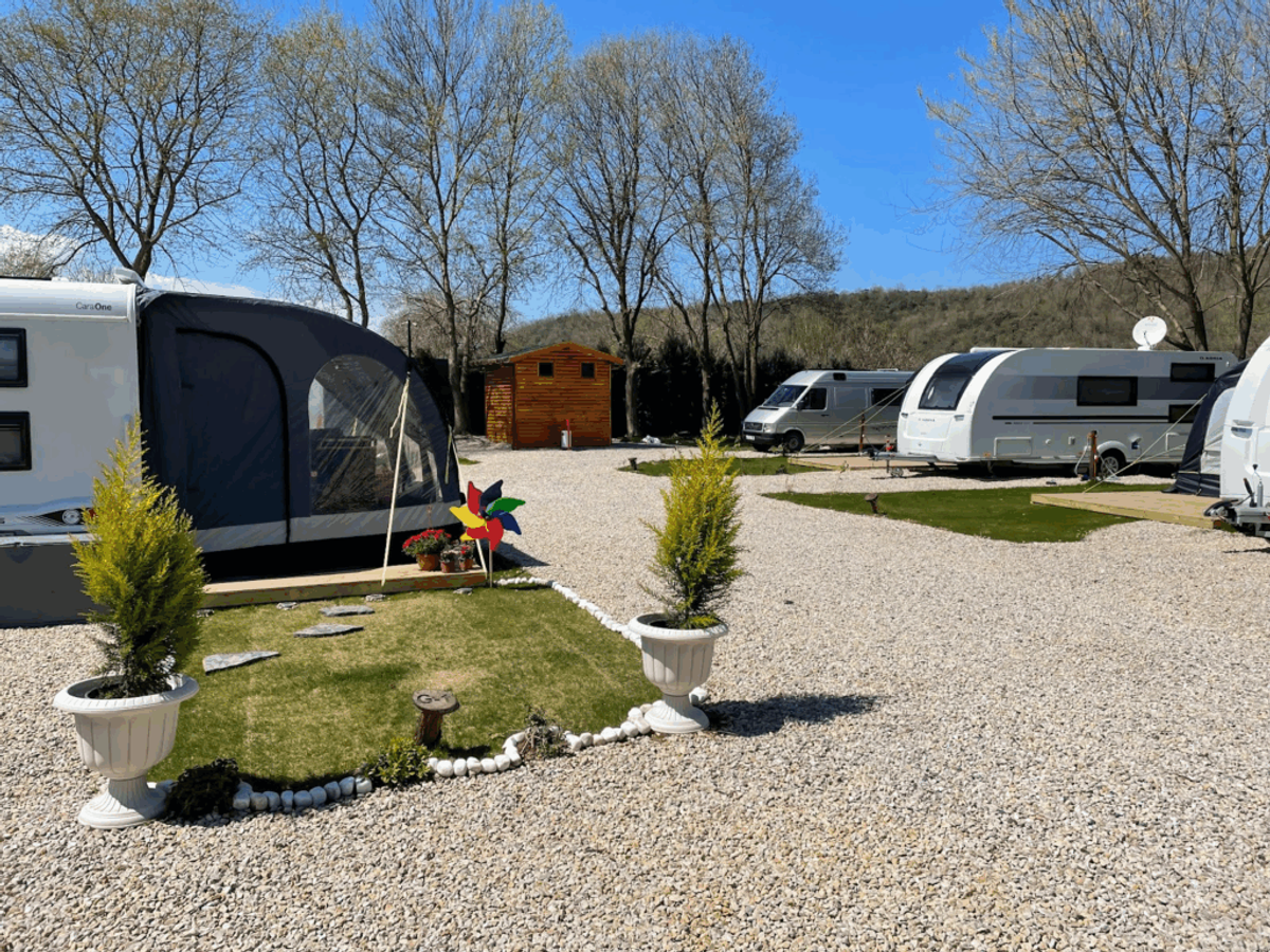 Well-maintained RV campground with multiple motorhomes parked on gravel sites, landscaped grass areas with potted plants, wooden facility building, and tree-lined hills in background under clear sky.