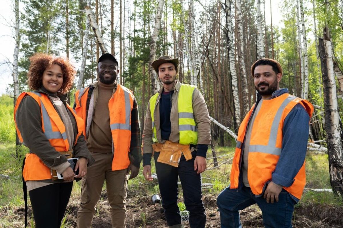 Four campground staff members wearing safety vests stand together on a forest path lined with birch trees, smiling at the camera.
