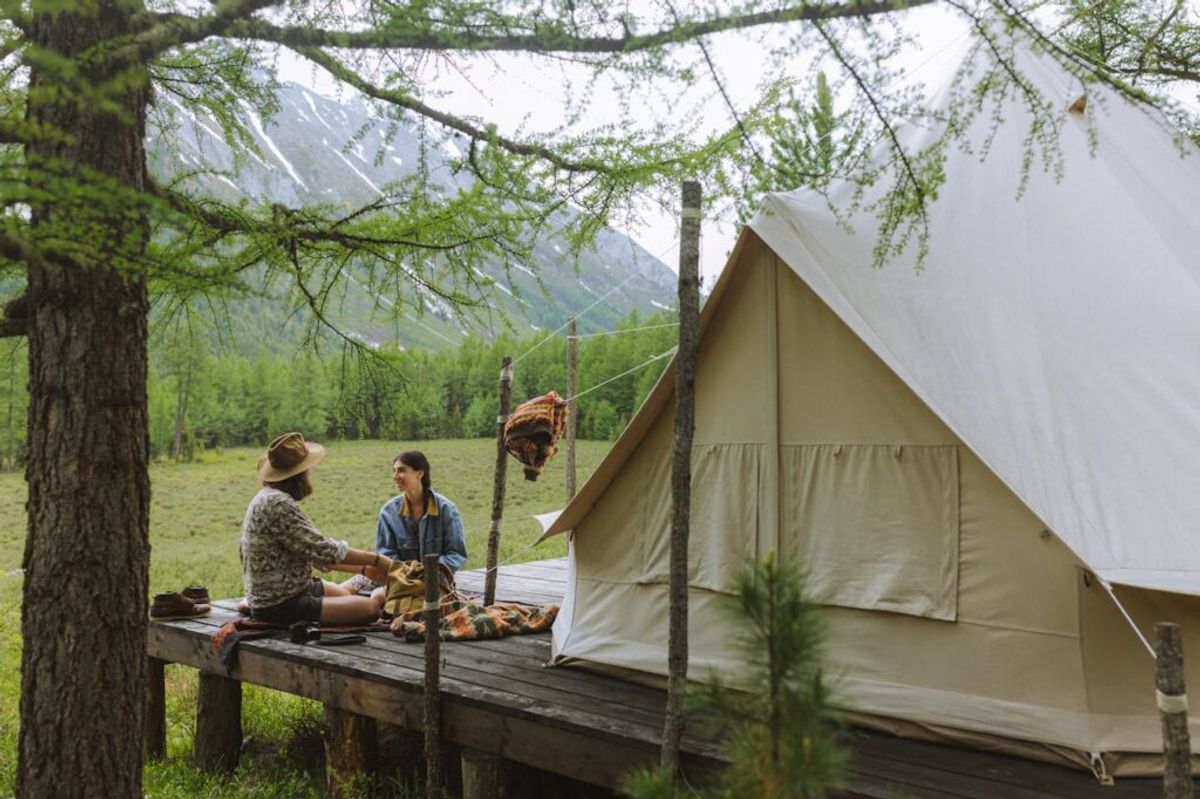 Two guests sharing a meal on a wooden deck outside a canvas glamping tent in a mountain valley setting.