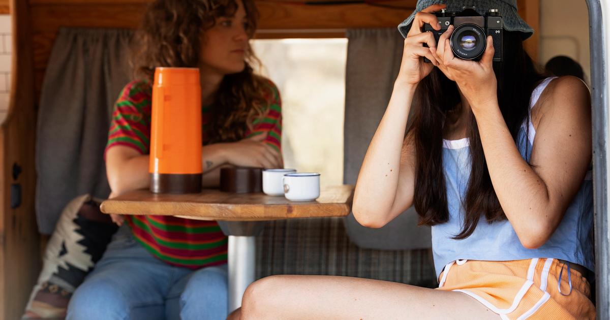 Two women inside an RV taking photos with a vintage camera, with an orange thermos and camping items on a wooden table.