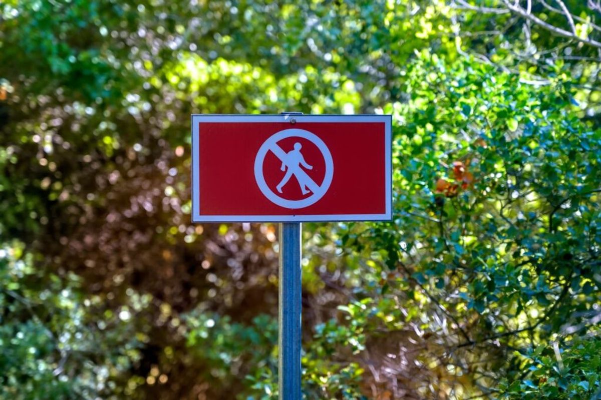 Red prohibition sign with white circle and figure, mounted on pole against blurred green foliage background.