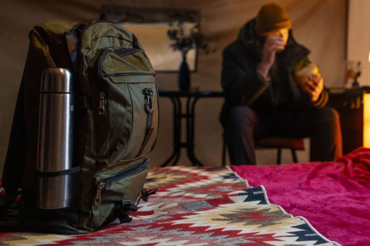 Backpack and thermos on bed with geometric blanket, person in background wearing hat and jacket in warm-lit room.