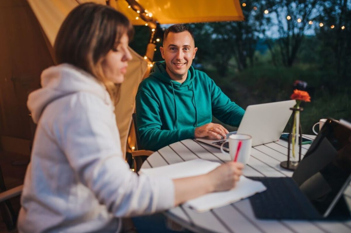 Two people working on laptops at an outdoor table under a yellow awning at dusk, with string lights and a natural backdrop.