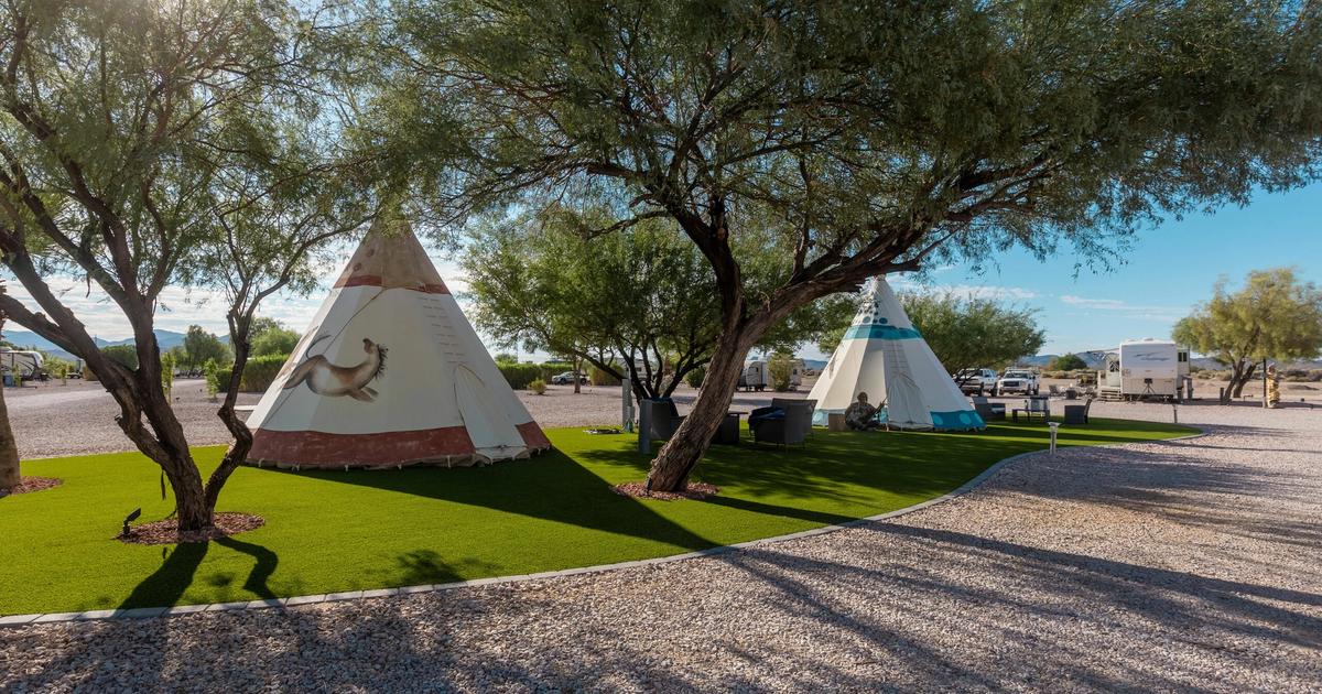 Two teepee-style accommodations on green artificial turf under large shade trees at an RV resort with gravel roads and RVs in background