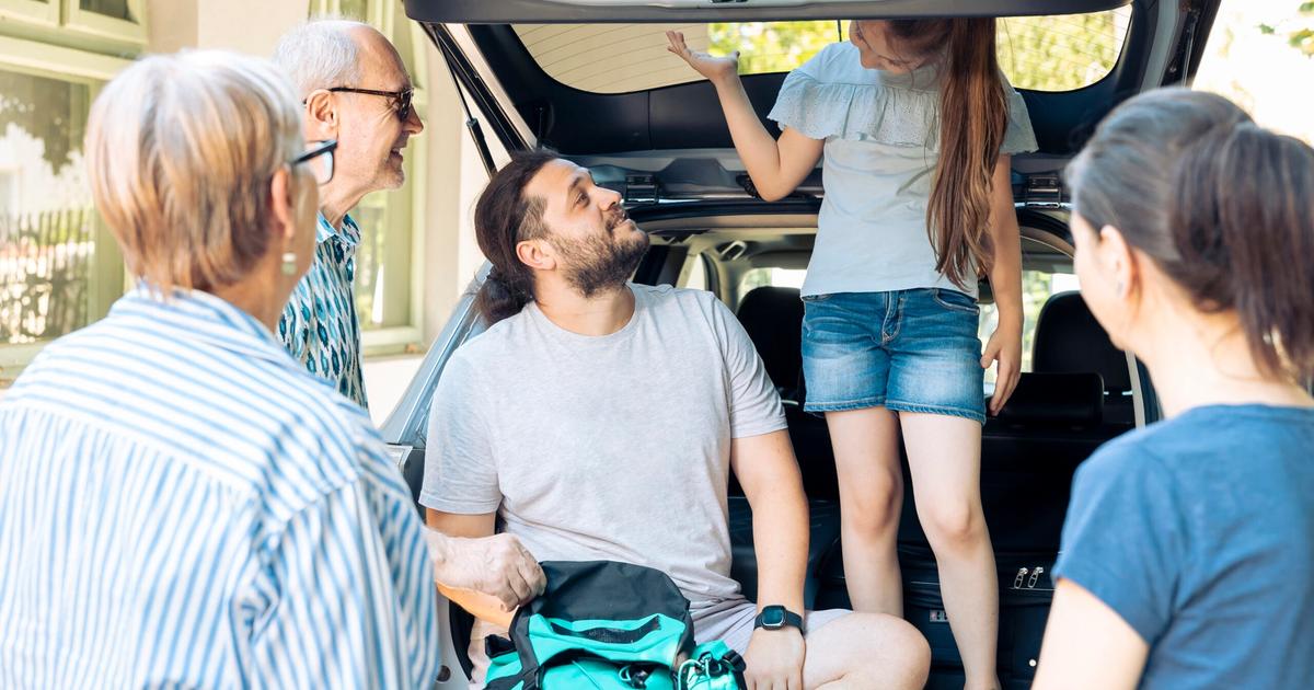 Family loading camping gear into car trunk while discussing travel plans outdoors.