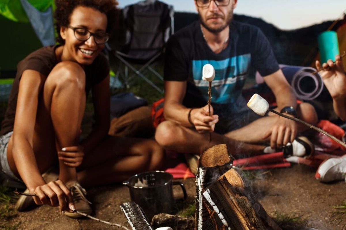 Group of friends roasting marshmallows over a campfire at dusk, sitting together in camping chairs outdoors.