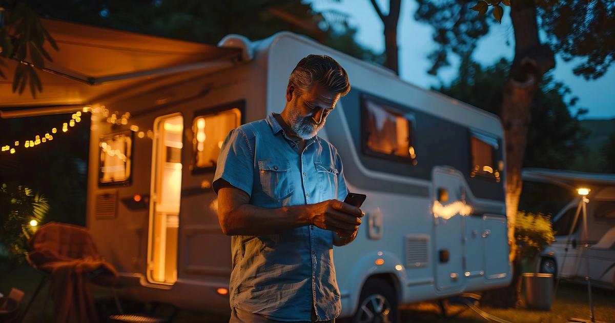 Bearded man in denim shirt checking smartphone outside illuminated RV at dusk in campground setting.