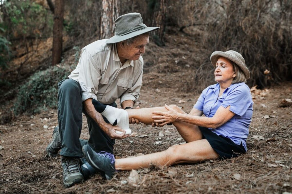 Man applying first aid bandage to woman's leg injury in forest setting, illustrating outdoor accident scenario.