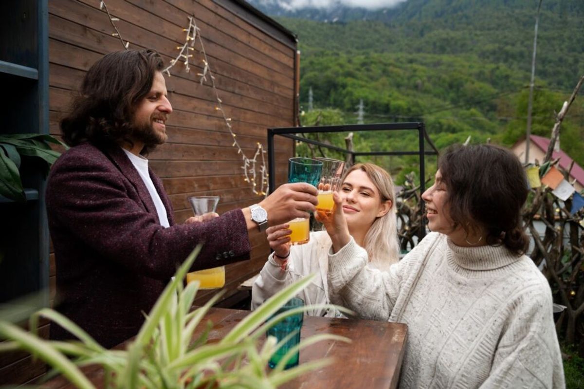Three guests toasting with glasses of juice at an outdoor venue with mountain views and wooden architecture in the background.