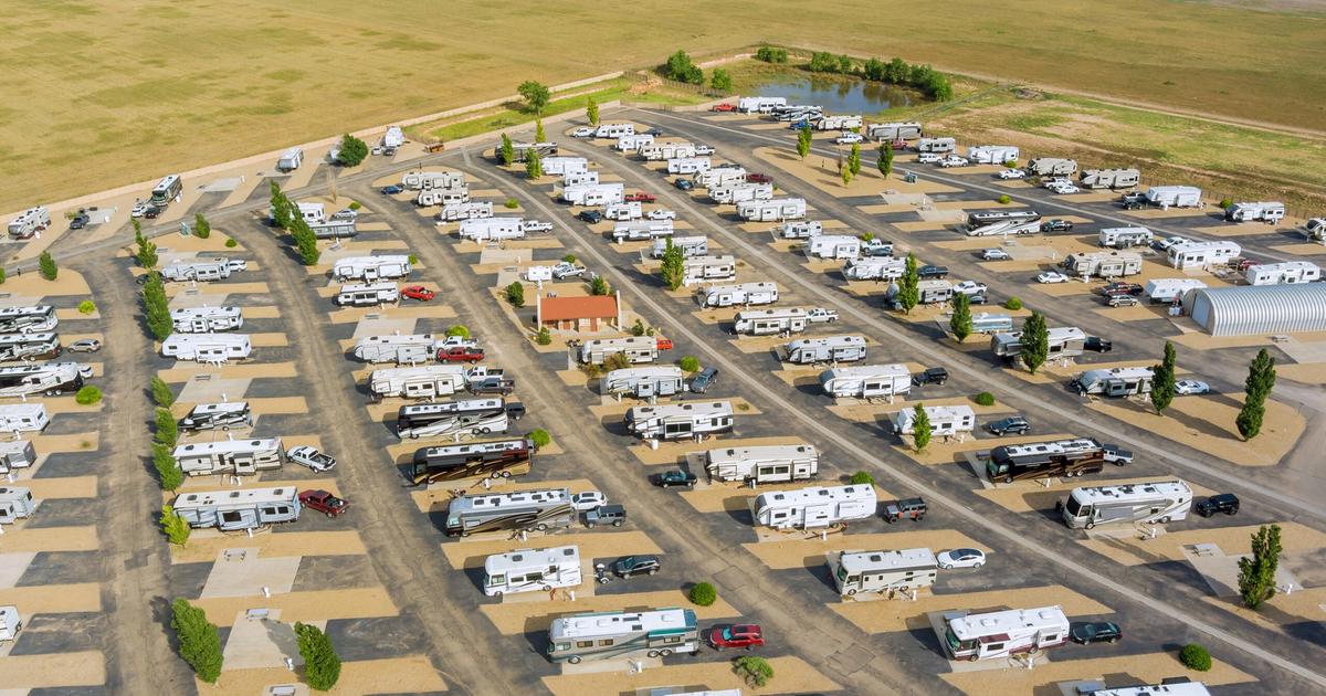Aerial view of a large RV and campground facility with organized rows of white recreational vehicles parked in numbered spaces, surrounded by green landscaping and rural terrain.