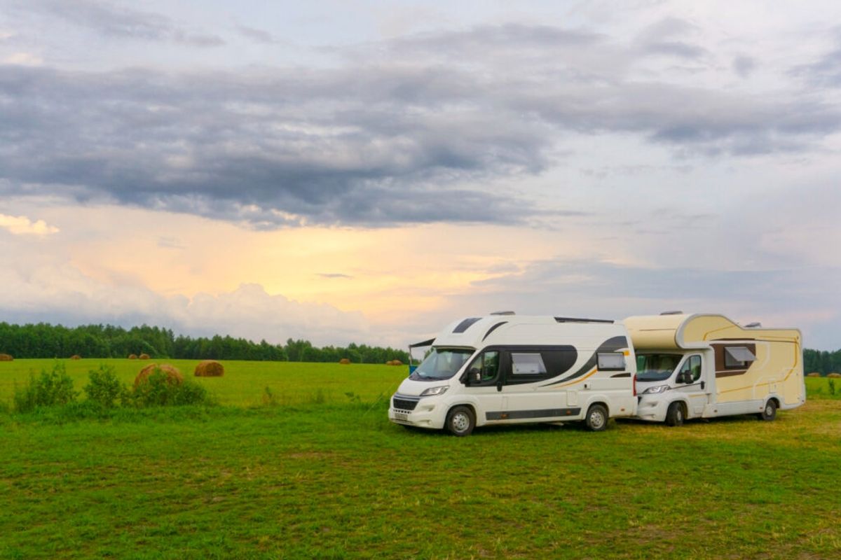 Two white RVs and a yellow camper parked in a green field with forest and cloudy sky in the background.