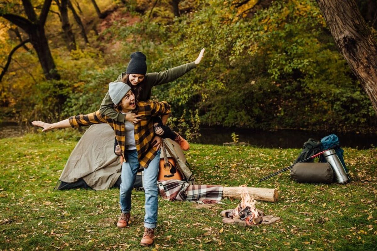 Two people enjoying campfire activities in autumn forest, one playing guitar while other gestures happily, with camping gear and fire nearby.