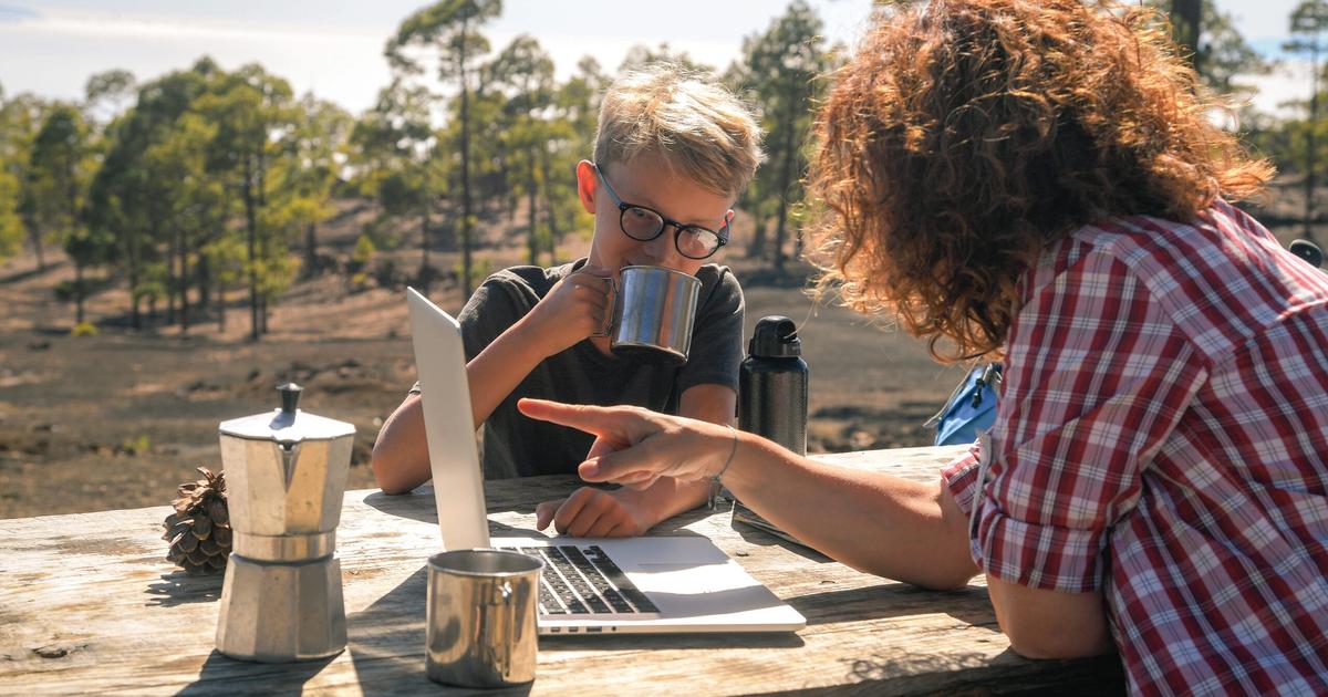 Two people working at a wooden table outdoors in a forest, reviewing laptop and documents with coffee, demonstrating remote campground management work.