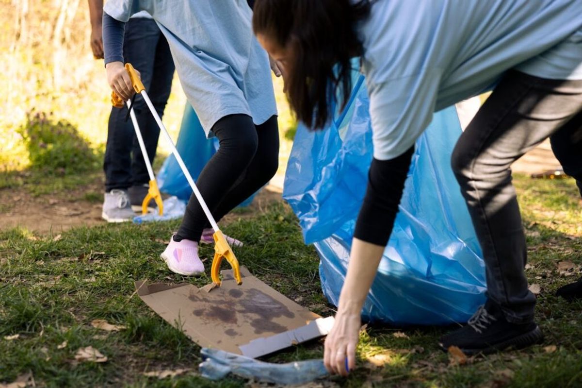 Volunteers in blue vests using grabber tools to collect trash into a bag during an outdoor cleanup activity on grass.